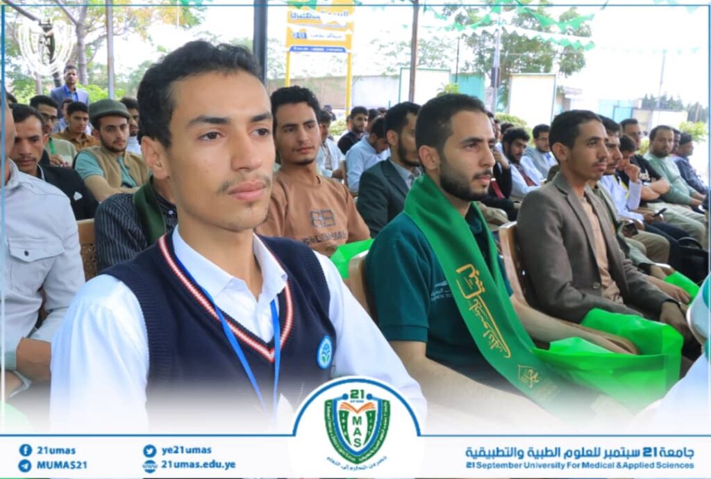 Group of students seated outdoors at a university ceremony; foreground man in white shirt and navy sweater vest with a lanyard, others behind him wearing casual clothes and a green scarf; banner with a university logo visible at the bottom.
