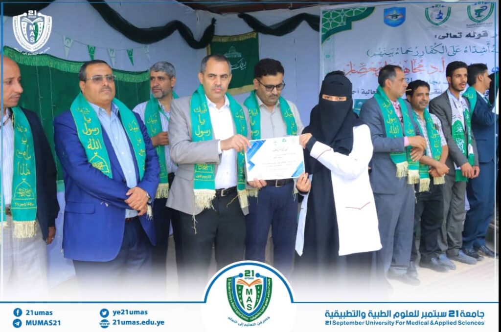 Group of men in suits and a woman in a black veil on a stage, handing a certificate during a university award ceremony with attendees wearing green sashes and banners in the background.