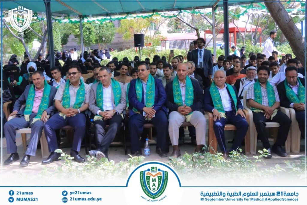 Group of men in suits wearing green sashes seated under a canopy at a university event, with attendees and greenery in the background.