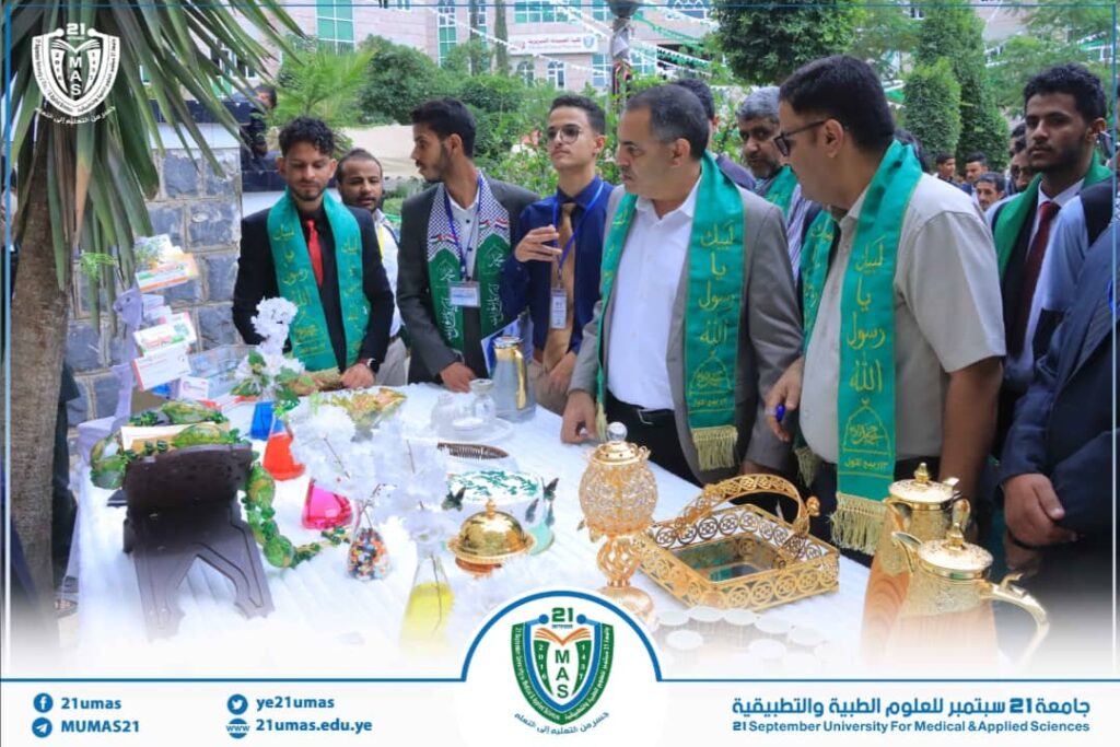 Group of men in suits with green ceremonial sashes stand around a table displaying ornate gold teapots, trays, and glassware at a university event.