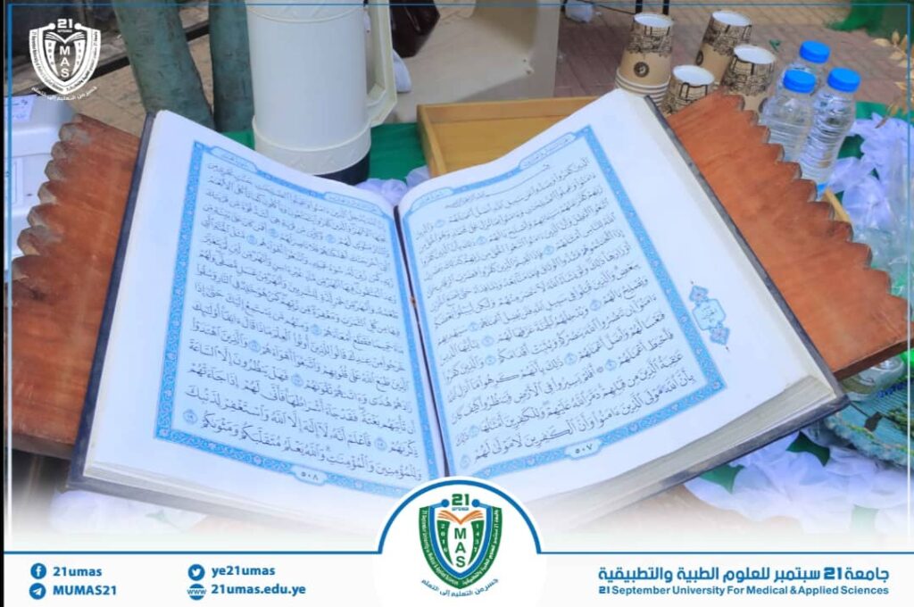 Open Qur'an on a decorative wooden stand during a university event, with branding at the bottom right and left.