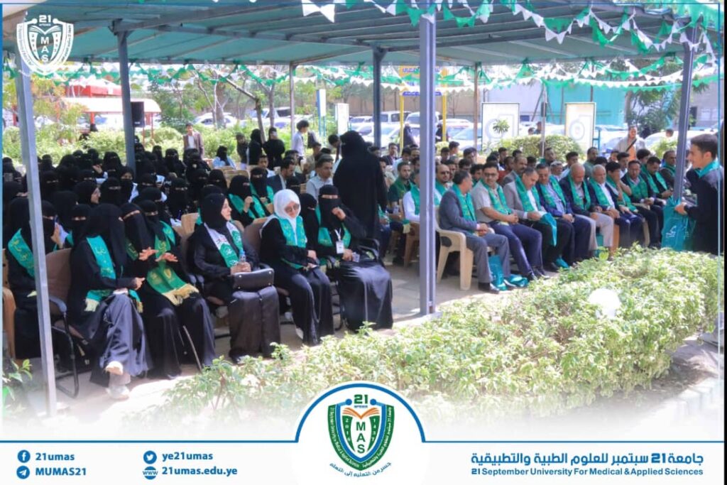 A large group of people seated in rows under a canopy at a university ceremony, many wearing green sashes and hijabs, with the campus crest visible at the front.