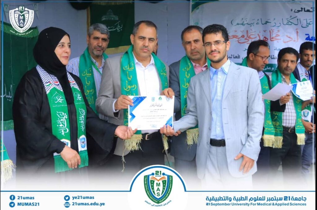 Group of adults on a stage during a university award ceremony; a man in a gray suit hands a certificate to a recipient in a light suit, with others wearing green sashes in the background.