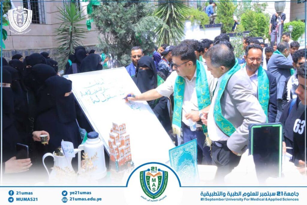 Group of students and faculty in front of a whiteboard at an outdoor university event, some wearing green sashes, while women in black abayas look on; university logo visible at bottom edge.