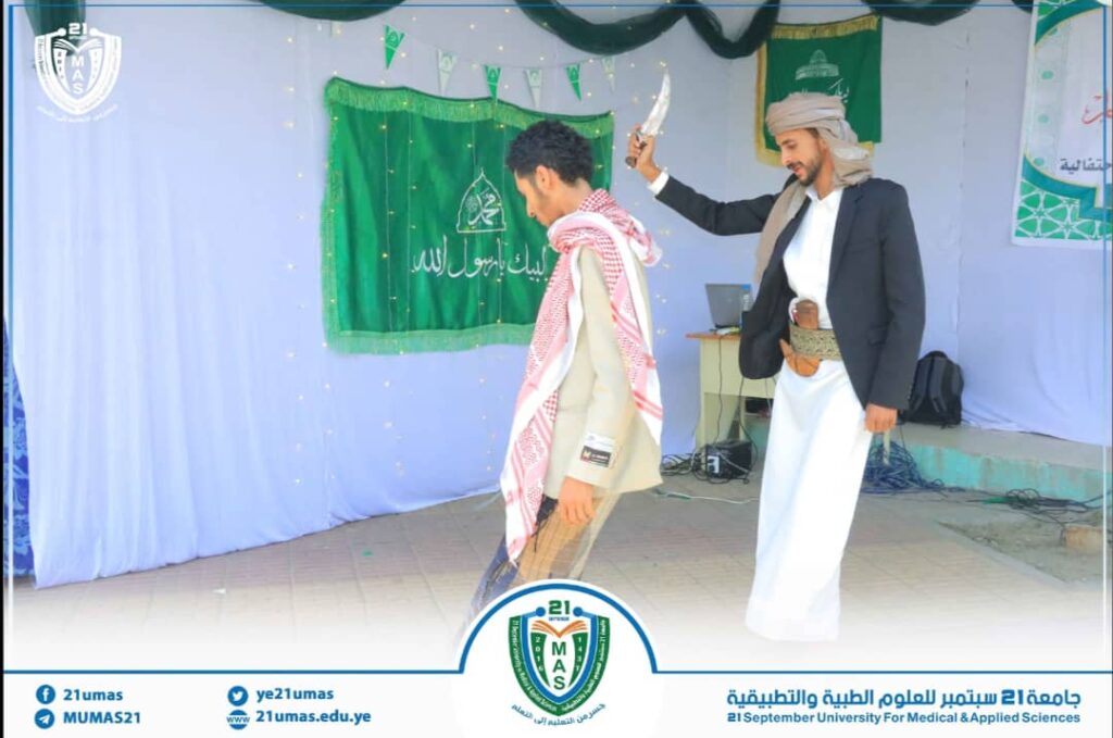 Two men in traditional attire perform on a university stage; one raises a knife toward the other during a ceremonial act, with banners and a crest in the background.
