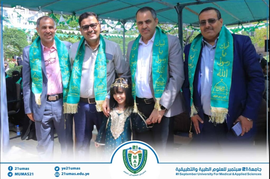 Four men in suits wearing teal ceremonial scarves stand with a smiling girl in a traditional dress under a tent at a university event.
