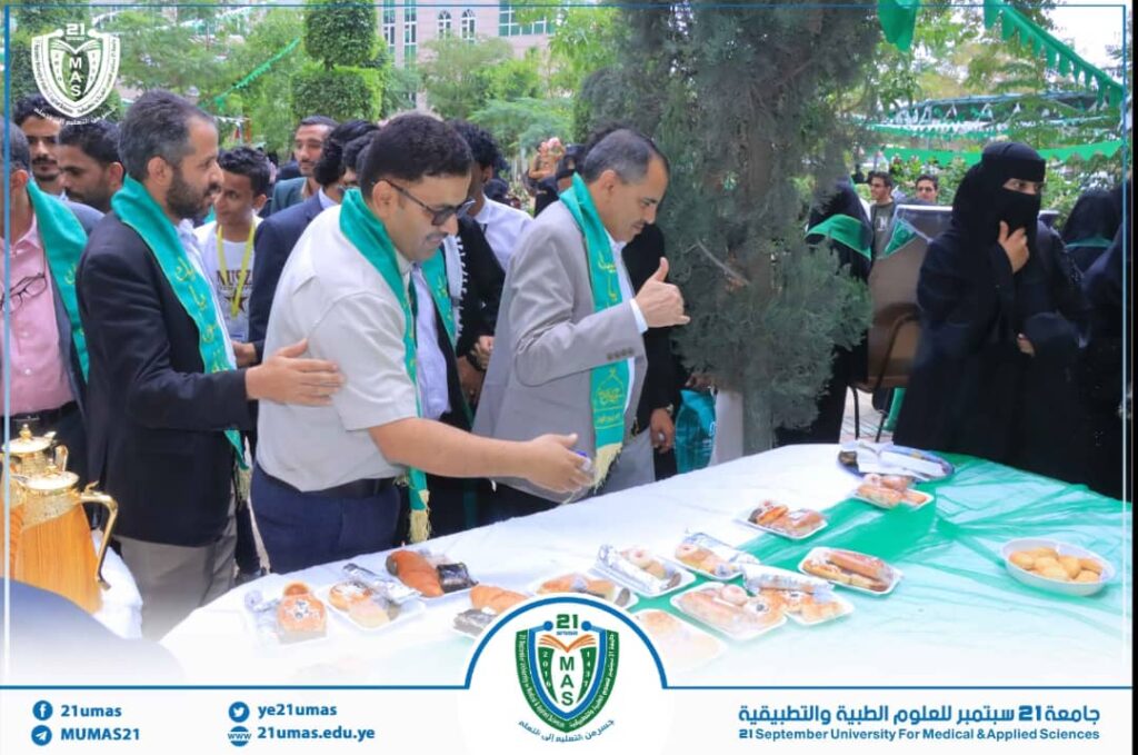People with green scarves line up to pick pastries from a long table at a university event; campus trees and a logo/banner are visible in the background, conveying a festive gathering.