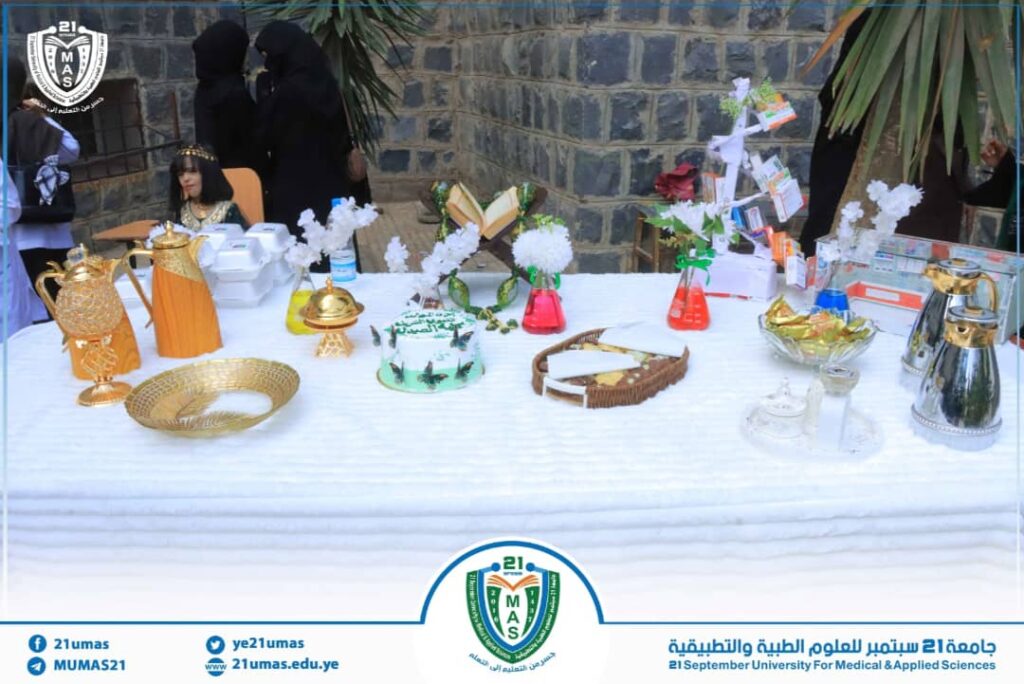 Table covered with a white cloth displays ornate gold teapots, a cake, bowls, and decorative white trees during a university event, with people in black abayas in the background.
