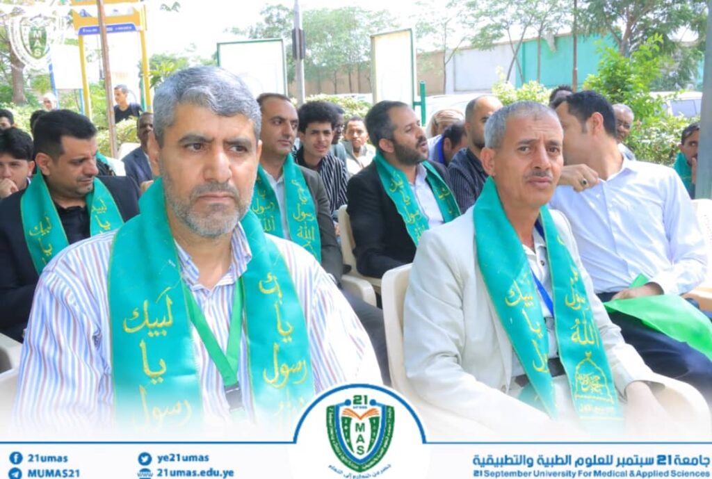 Group of men seated outdoors at a formal event, wearing teal-green sashes with Arabic script; banners and a university logo are visible along a decorative bottom border.