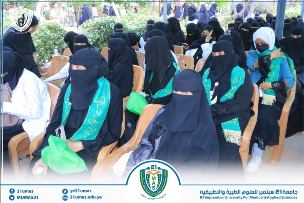 Group of women in black abayas and niqabs seated outdoors at a university event, wearing green sashes with Arabic text and a campus banner below.