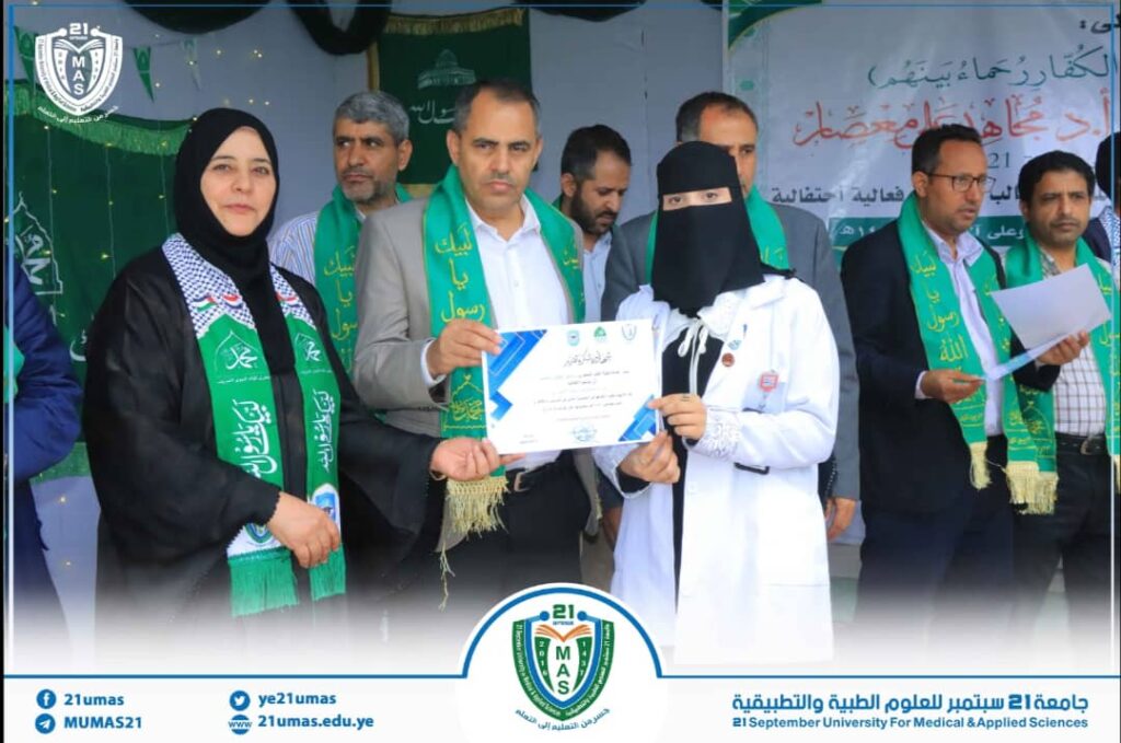 People on a university stage during a ceremony; a woman in a black hijab hands a certificate to a student in a white lab coat, both wearing green sashes, with a banner and logos in the background.