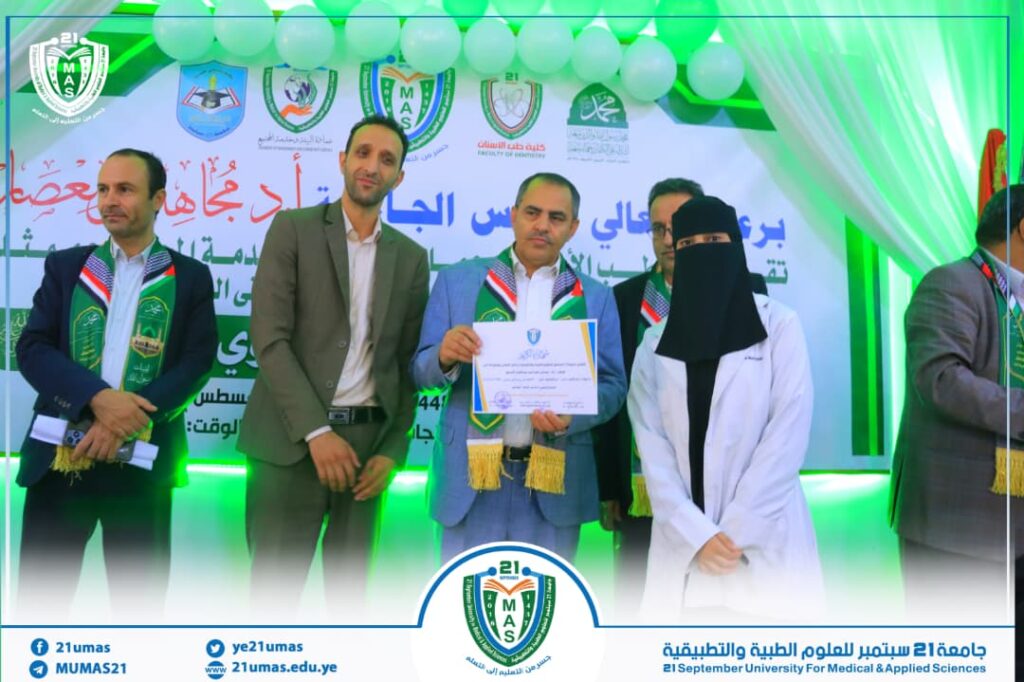 Ceremony on a stage with men and a veiled woman, one man holding a certificate, green decorations and university logos in the background