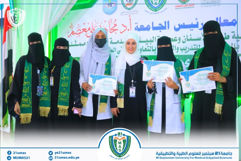 Five women in black abayas and hijabs stand on a stage, each holding certificates; they wear green sashes and face the camera, with a blue-green backdrop and university logos behind them.
