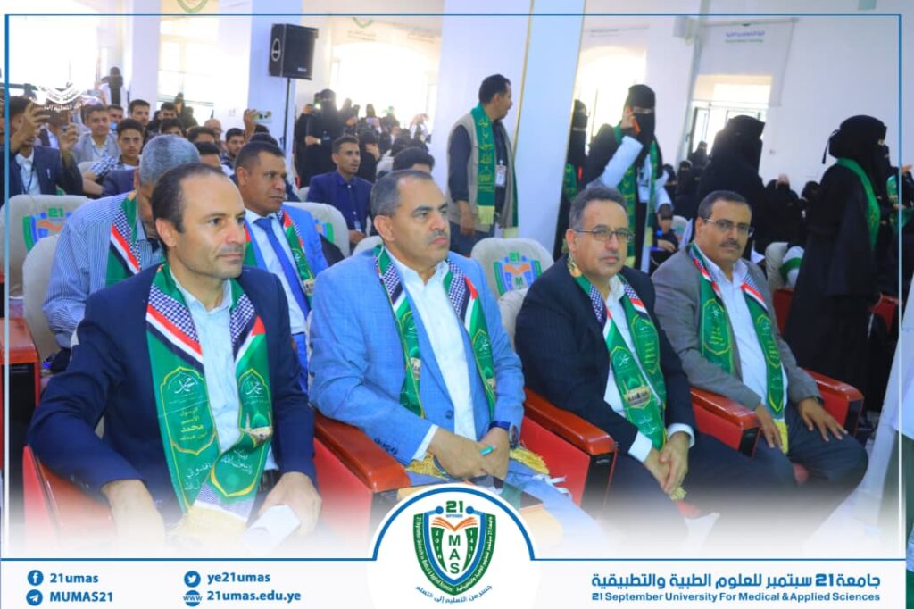 Group of men in suits with green ceremonial sashes seated in a row at a university event.