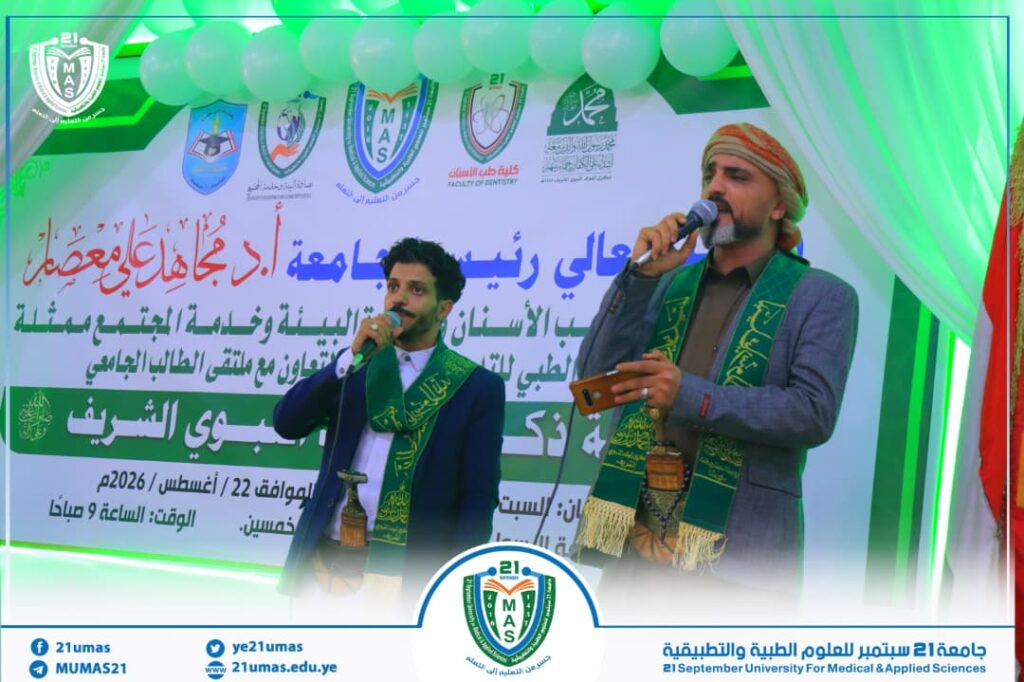 Two men in suits with green scarves speak into microphones on a decorated stage at a university event with Arabic banners and logos in the background on a green-themed setup.