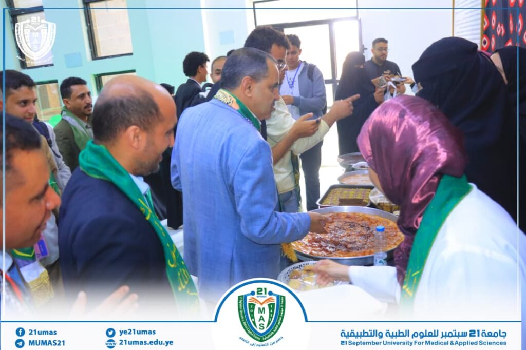 Group of people at a university event serving food from large bowls; a server wearing a purple hijab hands a dish to a man in a blue suit while others chat in the background.