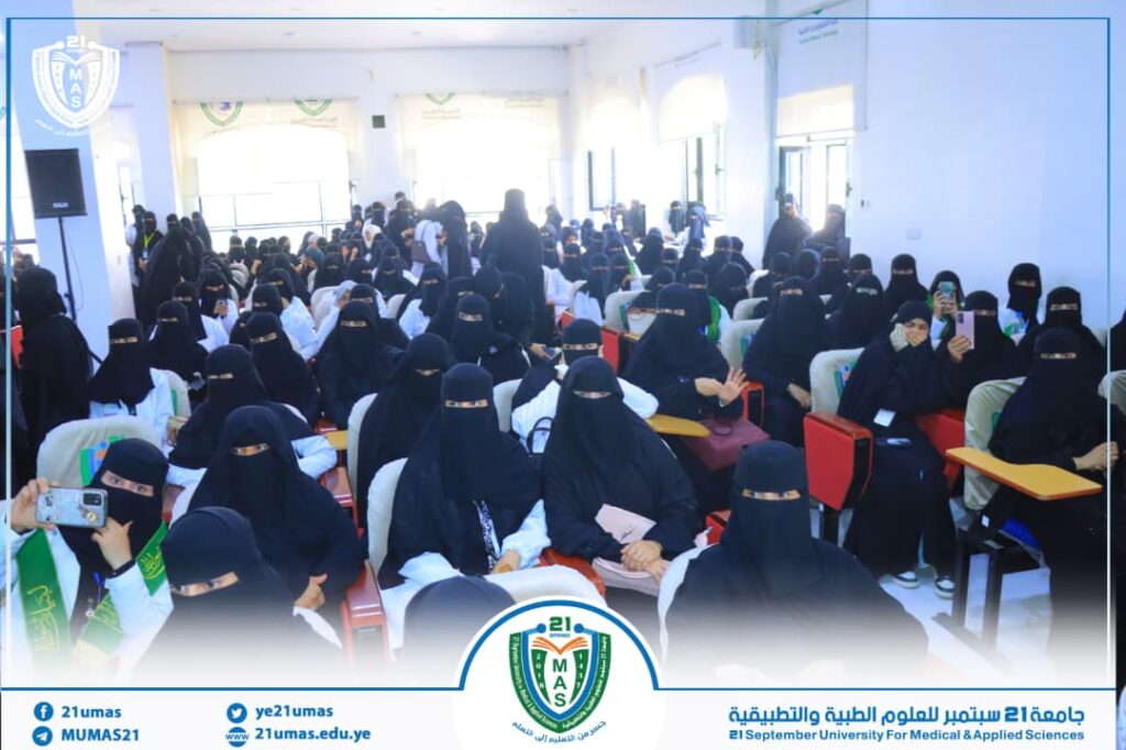 Wide view of a crowded auditorium filled with women in black niqabs, seated in rows for a university event; banners and logos are visible around the room.