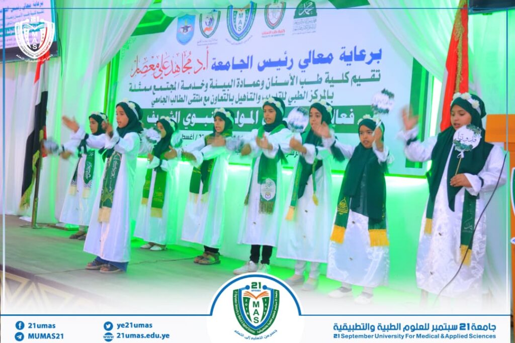 Group of girls in white dresses with green sashes performing a choreographed routine on a stage at a university event, with a green-lit backdrop and Arabic banner in the background.