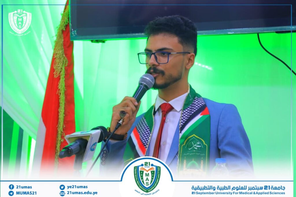 Young man in a blue suit and red tie speaks into a microphone on a green-lit stage, with the university crest visible in the background and on his sash.] 2012