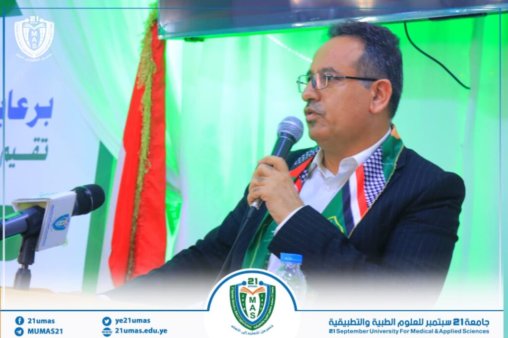 Male speaker in a dark suit and glasses, holding a microphone at a panel with a green backdrop and university banner behind him.