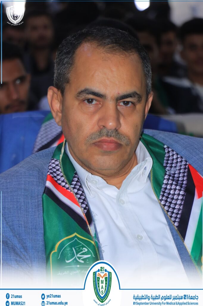 Close-up of a man at a university event, wearing a green ceremonial sash with a shield emblem; blurred audience in the background.