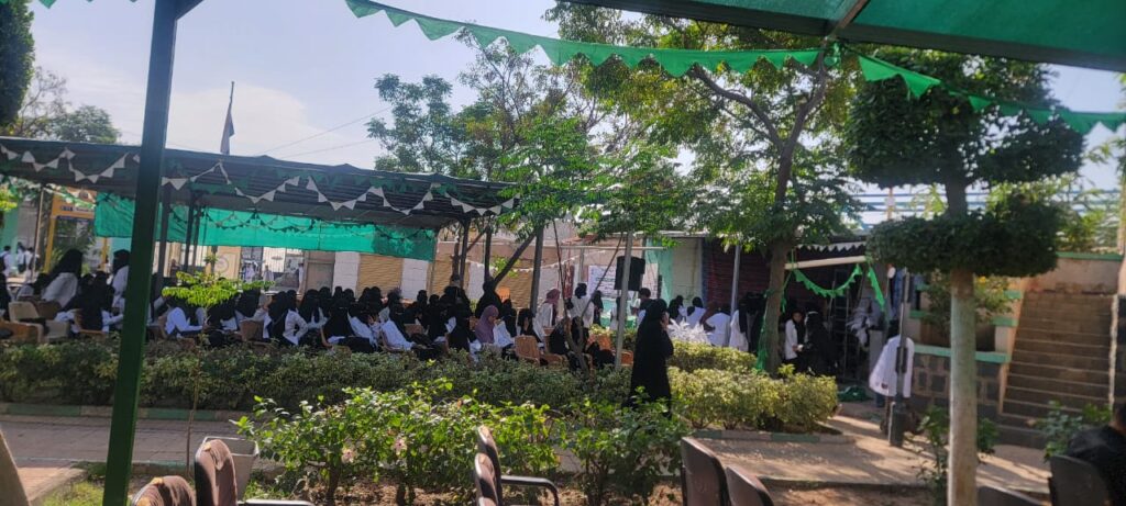 Students in white uniforms seated under green shade canopies at an outdoor school event under a sunny sky.