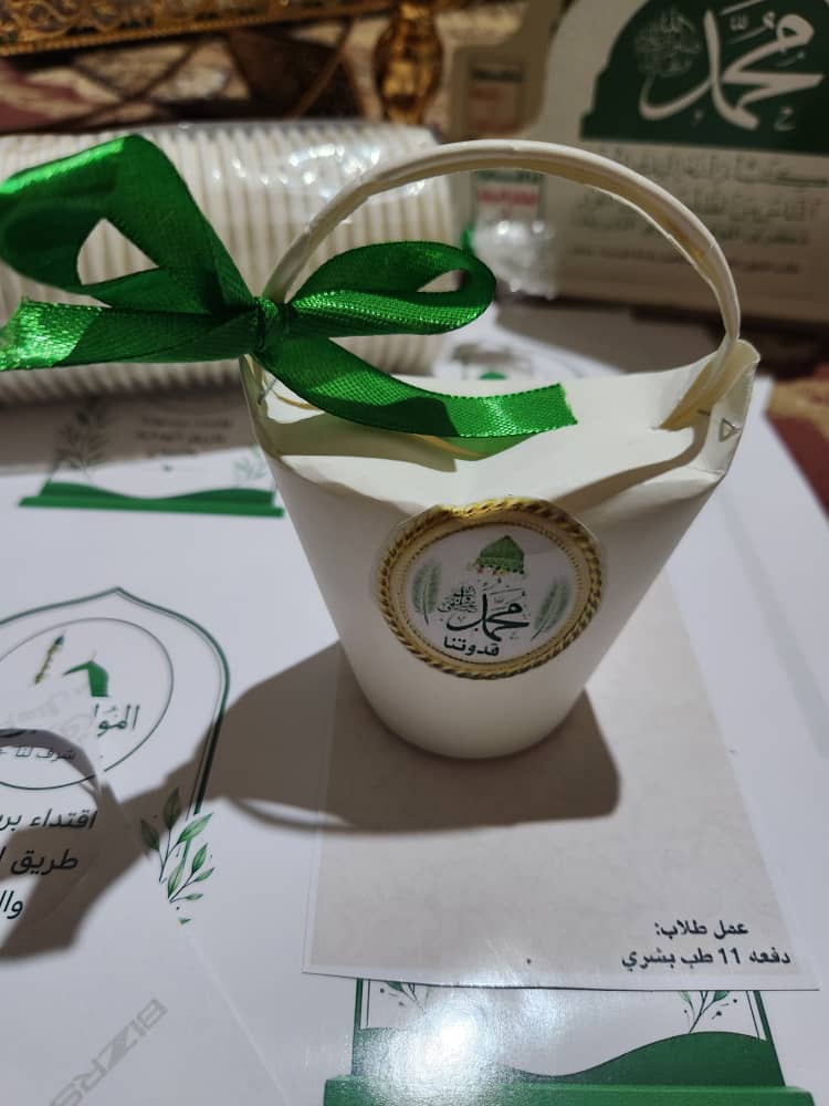 White paper gift bucket with a green ribbon and a gold-bordered seal, sitting on a table amid event cards (Arabic branding).