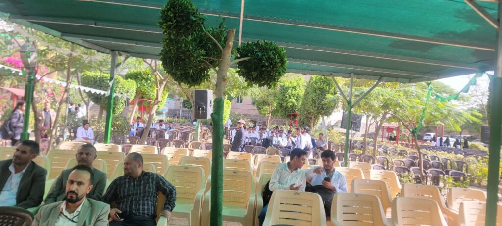 Outdoor event with rows of beige chairs under a green canopy; several men sit at the front left while two men converse on the right.