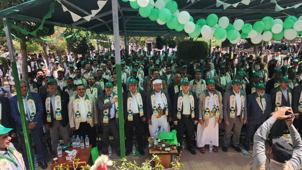 Large crowd of men under a green-and-white canopy at an outdoor ceremony, many wearing matching scarves and green caps, with balloons overhead.