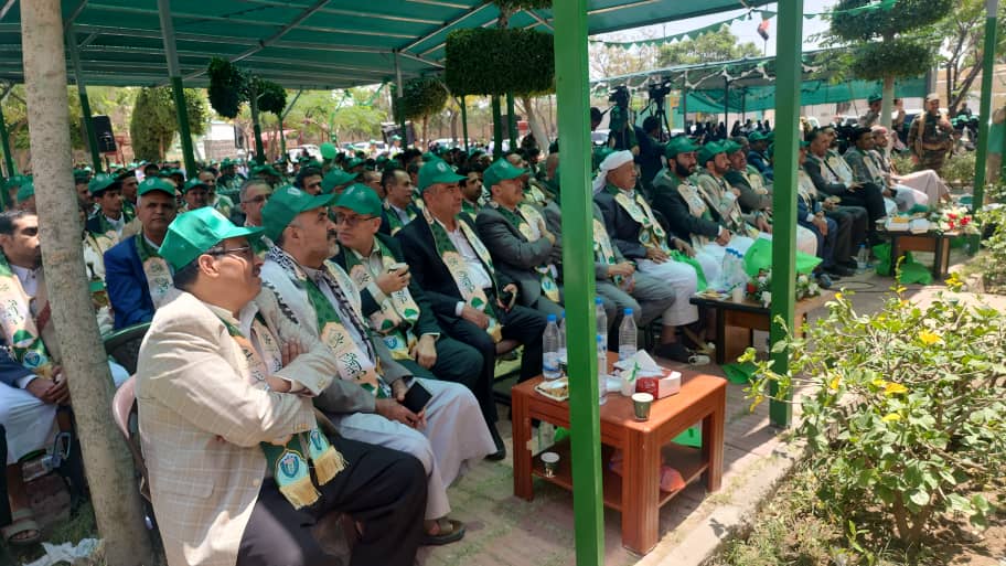 Large outdoor ceremony with men in green caps and scarves seated under a green canopy, listening attentively.