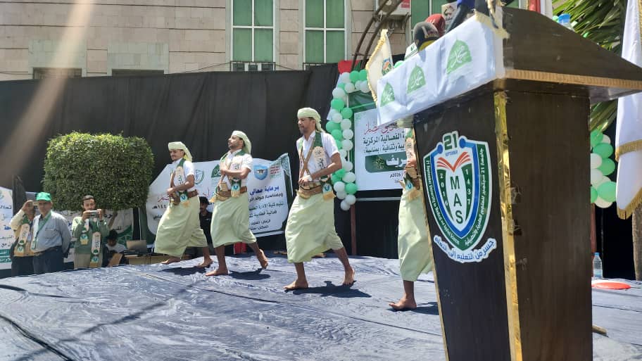 Four men in white traditional robes dance on an outdoor stage with banners in the background and a lectern nearby.