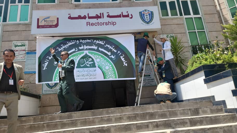 Group of men at the entrance of a university building labeled 'Rectorate' (الرئاسة الجامعة) hanging a large green and white banner on the stairs; a man on a ladder and others assisting while a speaker addresses from the steps.