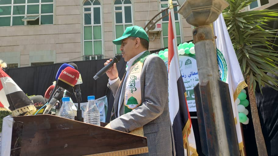 Man in a gray suit and green cap speaking into a microphone at an outdoor podium during a ceremony, with flags and balloons in the background.