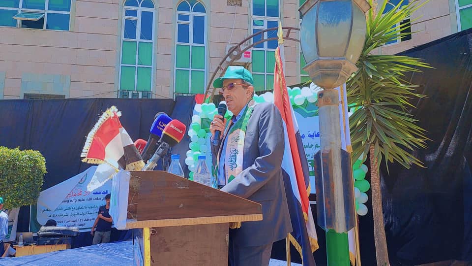 Man in a suit and teal cap speaks into a microphone at a wooden podium during an outdoor ceremony, with multiple microphones, a flag, green and white balloons, and a banner in the background.