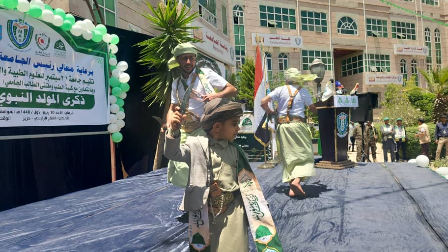 Young boy in traditional robes and headscarf holding a ceremonial sword on a stage during an outdoor ceremony, with banners and other children in similar outfits behind him.