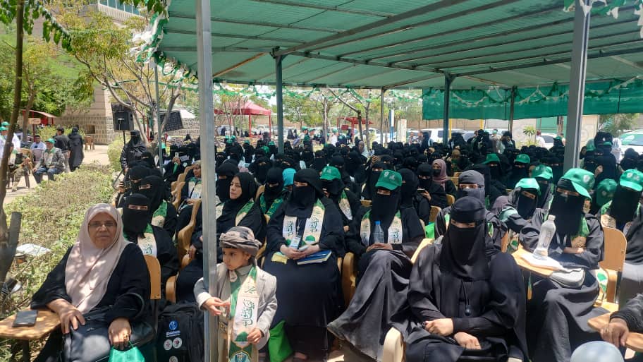 Group of women in black abayas and niqabs seated under a green canopy at an outdoor event, some wearing green caps.