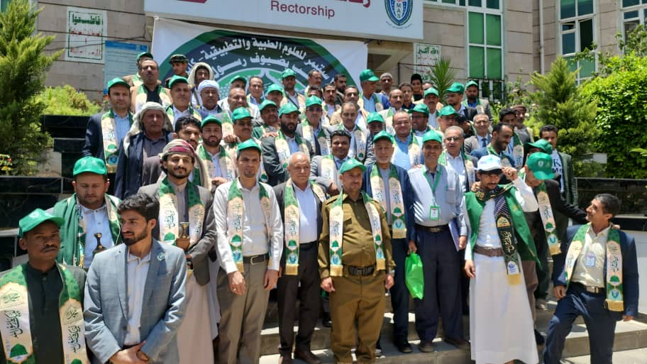 Group of men in green hats and sashes posing on steps outside a university building during a ceremony.