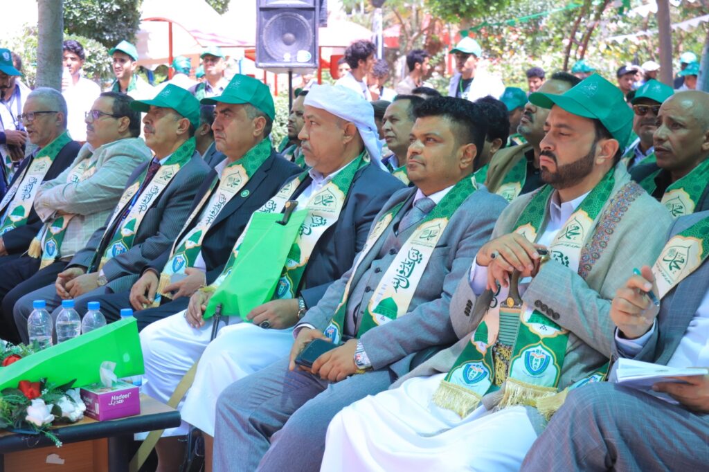 Group of formally dressed men seated outdoors at a ceremony, wearing green scarves and caps with badges while others stand in background.