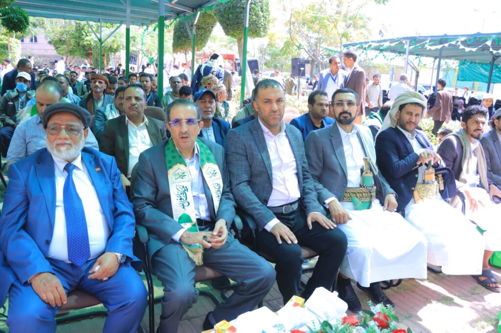 Front row of men in suits and traditional robes at an outdoor ceremony, with a large crowd under a canopy behind them.