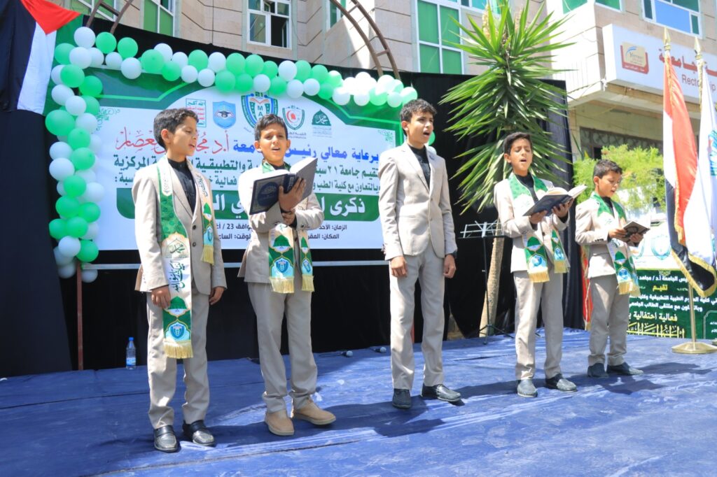 Five boys in beige suits with green ceremonial sashes sing on a stage, each holding a book during a school event, with a banner and green balloons behind them.