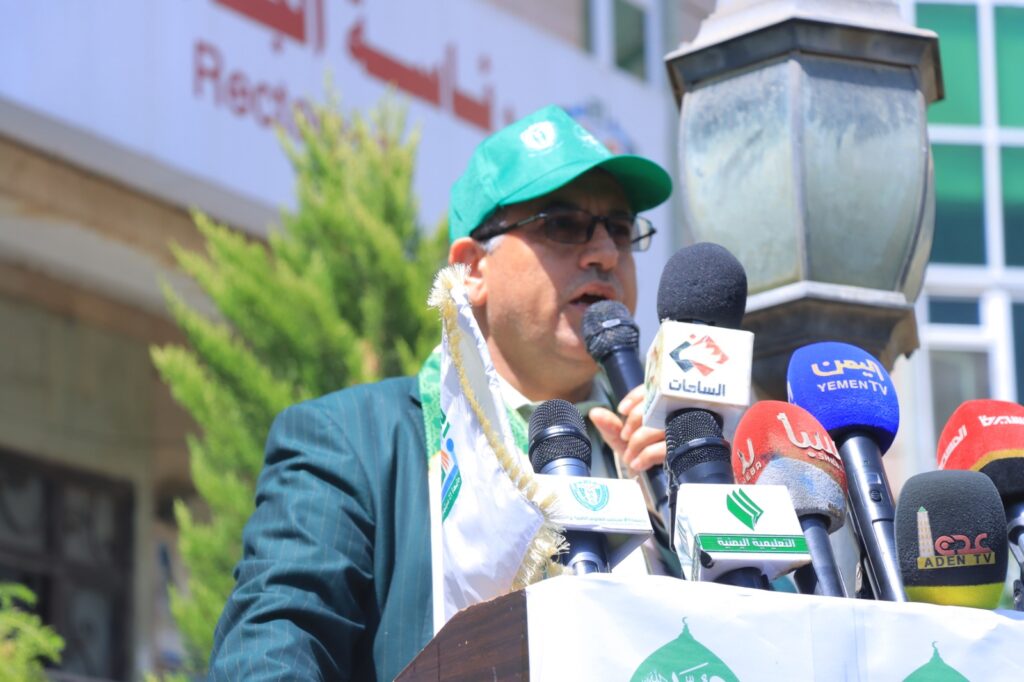 Man in a green cap and glasses speaks into a microphone at a press conference, with multiple TV microphones in front.
