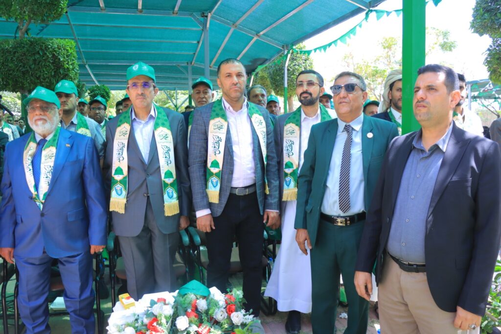 Group of men in suits wearing green scarves and caps at an outdoor ceremony under a teal canopy, with floral arrangements in front.