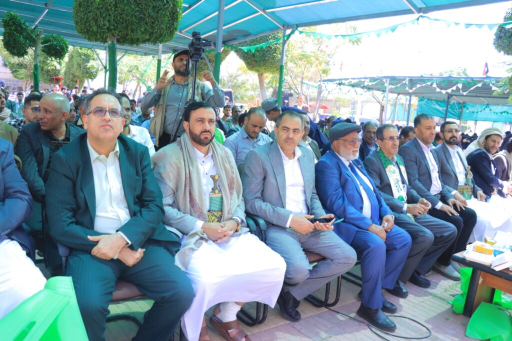 Group of men in suits seated at an outdoor ceremony under blue canopies, with a cameraman on a tripod behind them taking video.