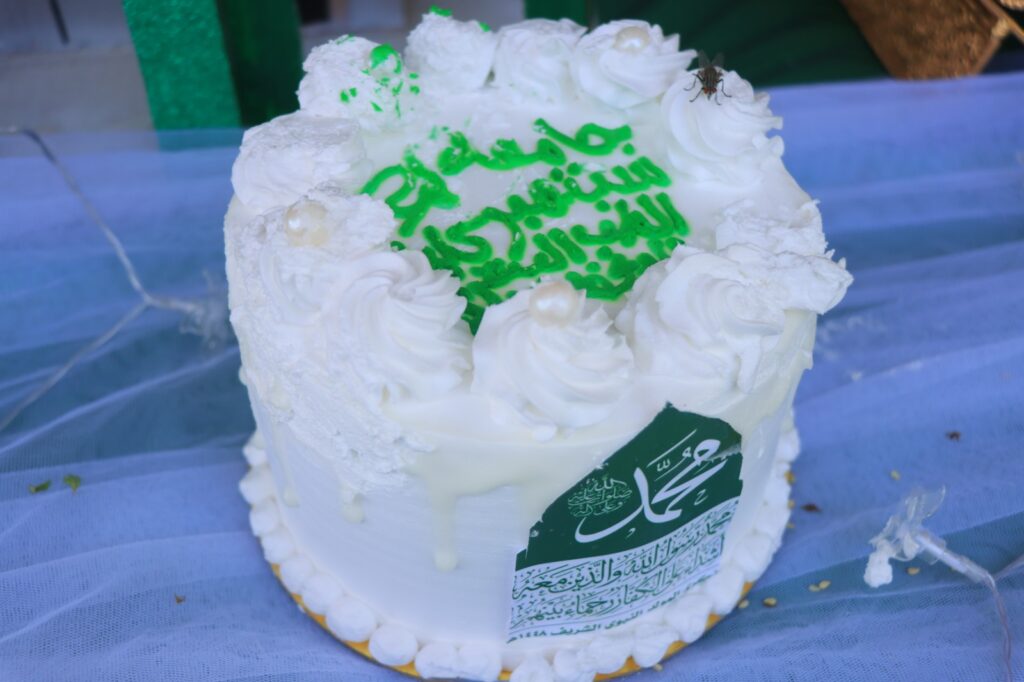 White frosted cake with green Arabic writing, white frosting rosettes, and a green label on the side.