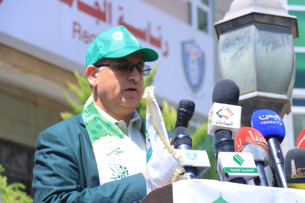 Mid-aged man in a green cap and suit speaks at a podium with multiple microphones during a press conference.