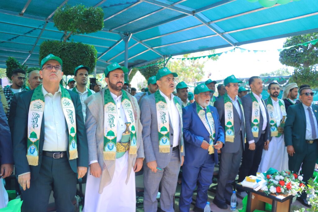 Group of men in suits wearing green scarves and caps, standing at an outdoor ceremony under a blue canopy.