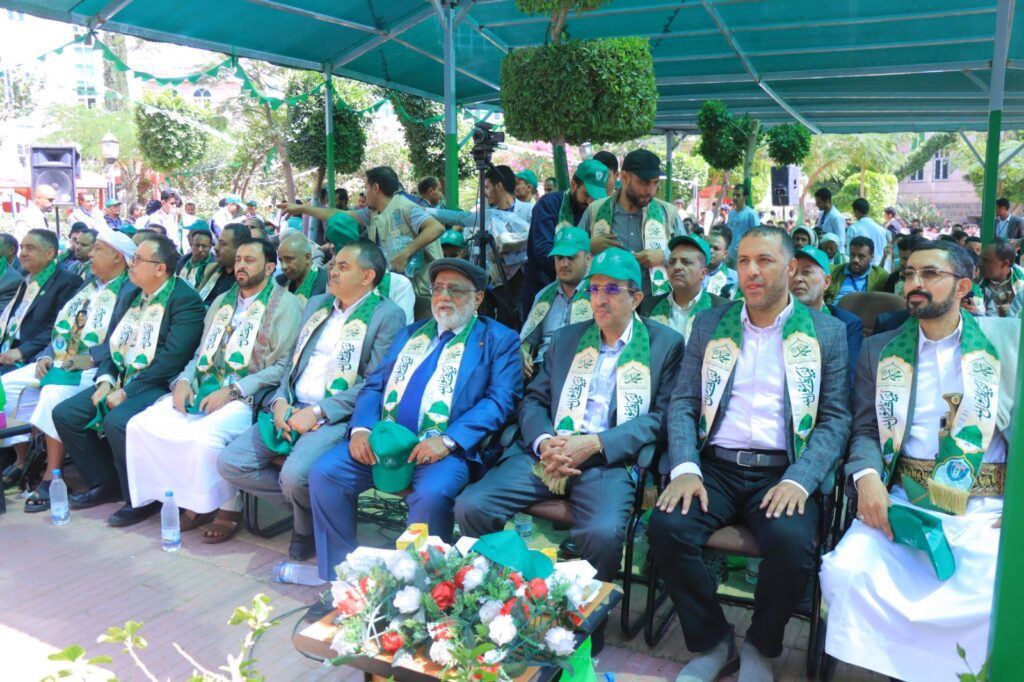Group of men in suits seated in an outdoor ceremony under a teal canopy, wearing green scarves and hats as attendees look on.
