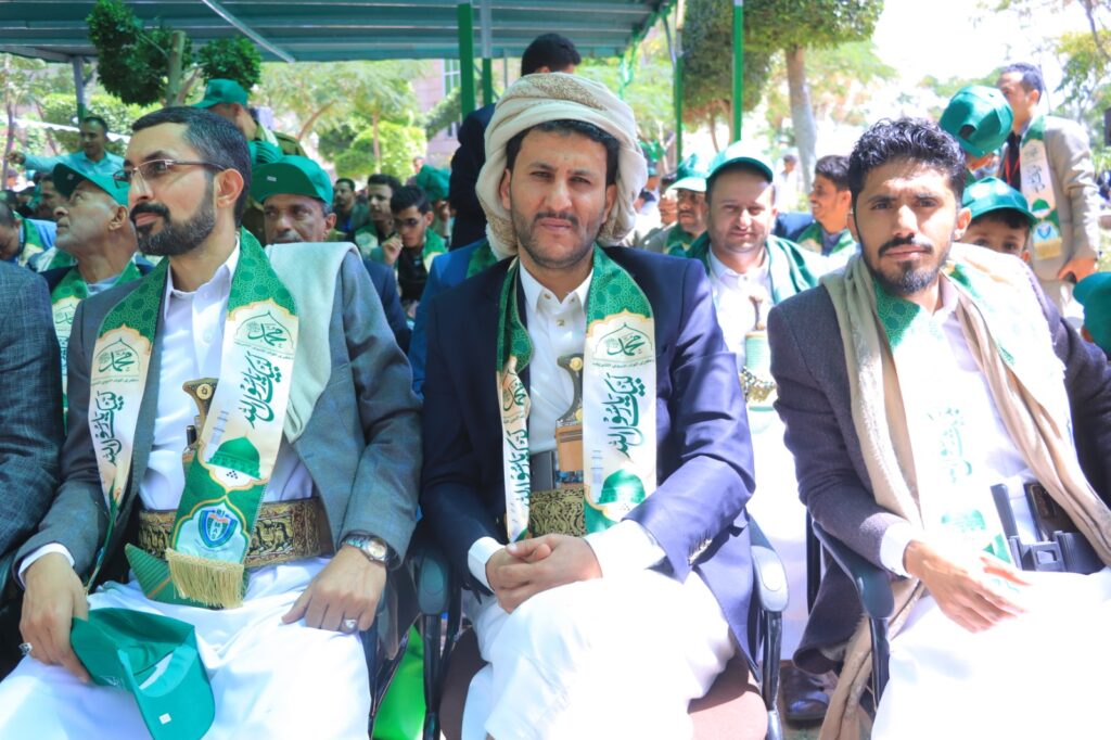 Group of men seated outdoors at a green-themed event, wearing white robes, suits, and green scarves with Arabic inscriptions.