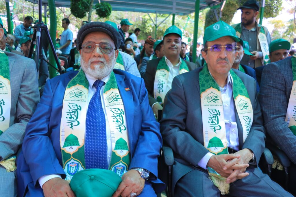 Group of men in formal attire wearing green sashes at an outdoor ceremony.