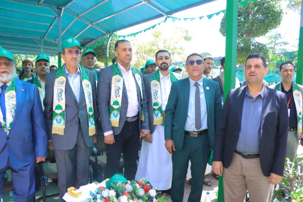 Group of men in suits with green sashes and hats at an outdoor ceremony under a teal canopy.